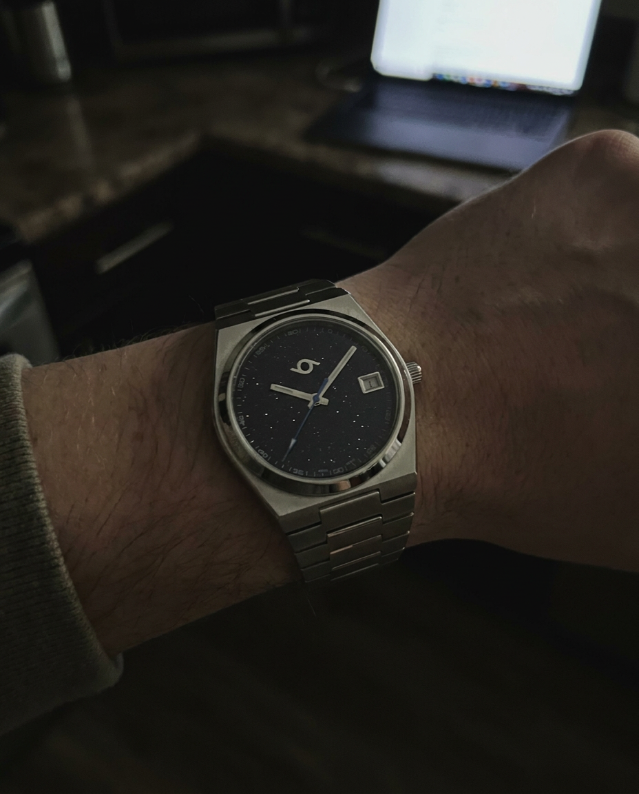 A silver-toned metal wristwatch with a dark blue dial and date window, worn on a person's wrist in a dim indoor setting with a blurred laptop in the background.