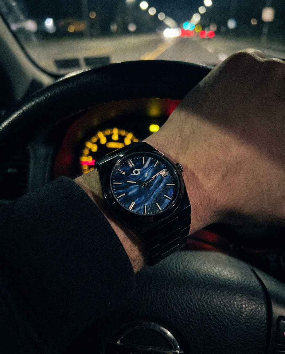 A wrist wearing a black watch with a textured blue dial, shown while a hand rests on a car steering wheel at night.